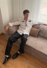Jeune homme portant une chemise blanche boutonnée et un pantalon noir, assis détendu sur un canapé beige dans une pièce ensoleillée.