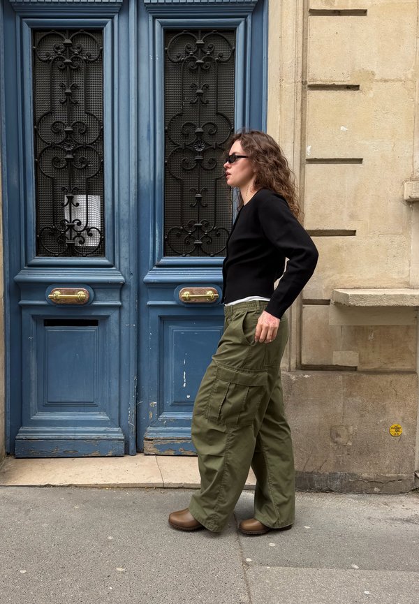 Frau mit Sonnenbrille, schwarzem Top und olivfarbenen Cargohosen geht an einer blauen Doppeltür mit verzierten schwarzen Metallgittern an einer beigen Steinwand vorbei.