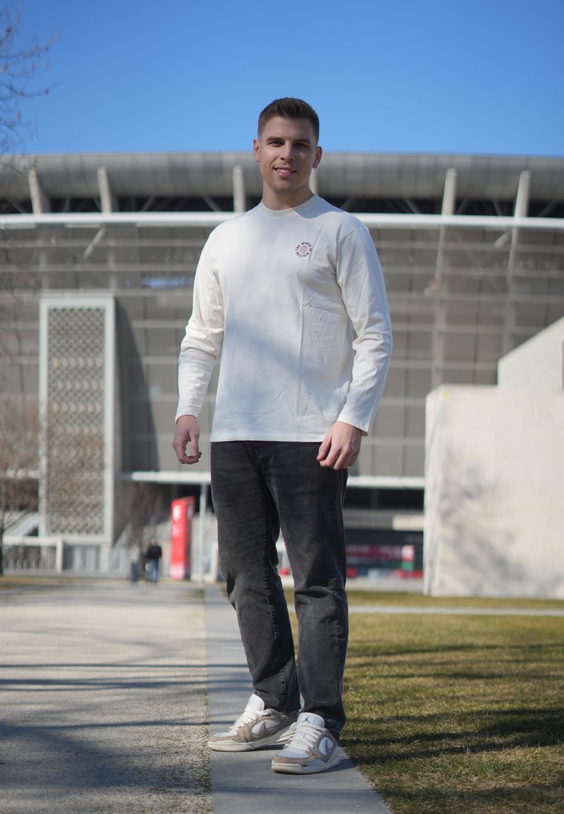 Jeune homme portant une chemise blanche à manches longues et un jean noir, debout sur le trottoir avec un grand bâtiment et un ciel bleu en arrière-plan.