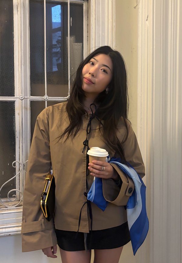 Jeune femme aux longs cheveux bruns, portant une veste beige et une jupe noire, tenant une tasse de café et une écharpe bleue à l'intérieur près d'une fenêtre.