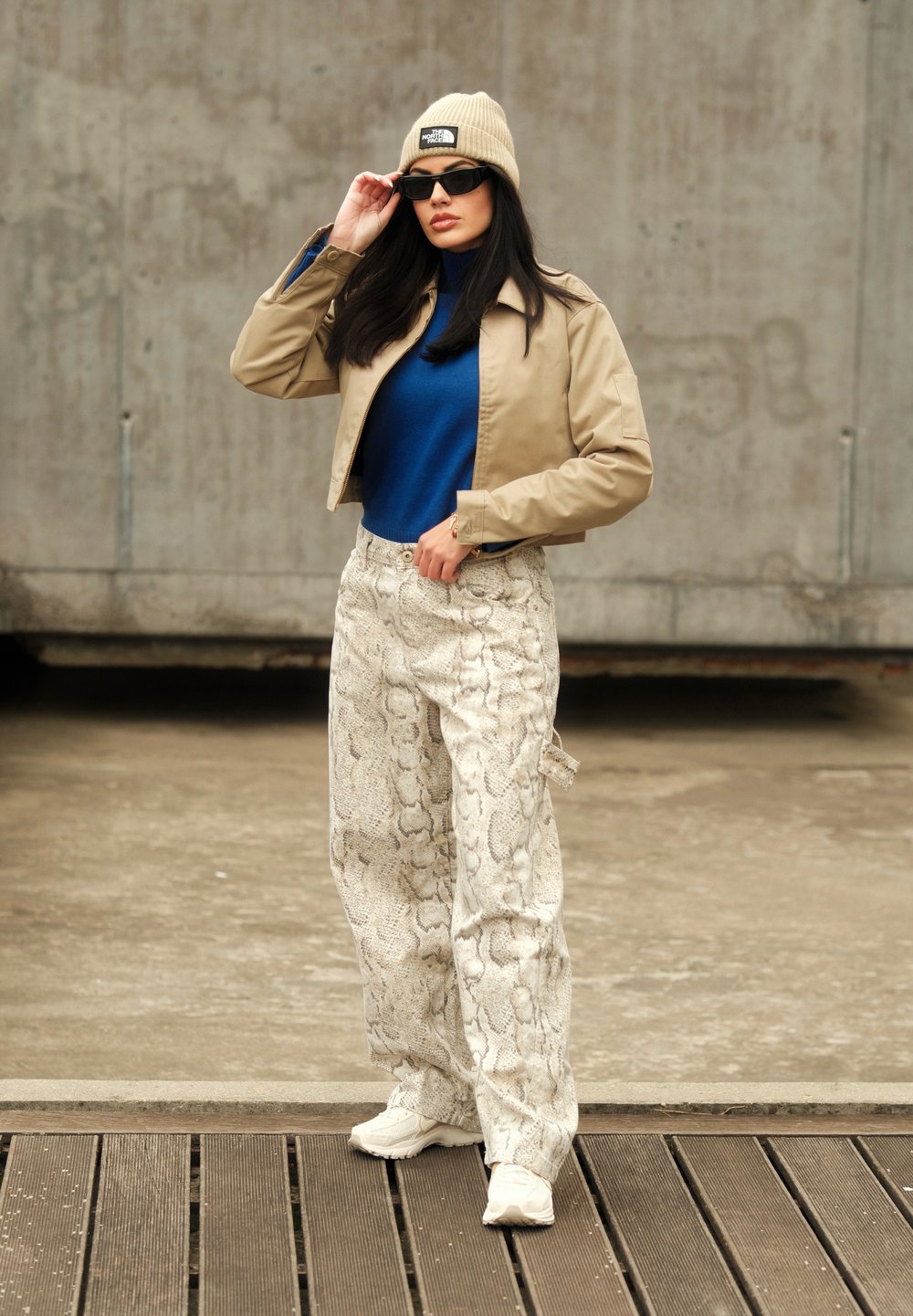 Μπεζ κοντό μπουφάν, μπλε πουλόβερ με ψηλό γιακά, λευκά παντελόνια με σχέδιο από φίδι, μπεζ σκουφάκι και γυαλιά ηλίου;αθλητικά παπούτσια ολοκληρώνουν την εμφάνιση.