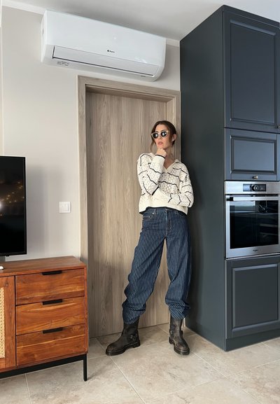 Suéter grueso de crema con líneas negras, pantalones de rayas azul marino de tiro alto y botas negras de tobillo. De pie en un ambiente de cocina moderna.