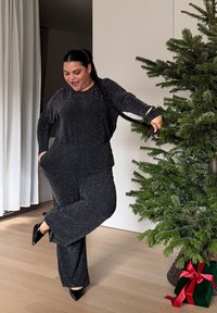 Sort, glitrende loungewear-sæt med en løstsiddende top og vidde bukser. Modellen bærer sorte høje hæle og står nær et grønt juletræ.