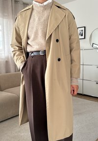 Henkilö, joka on pukeutunut beigeen trench-coattiin, beigeen neuleeseen valkoisen paidan päällä, ruskeisiin housuihin ja mustaan vyöhön, seisoo sisätiloissa käsi taskussa.