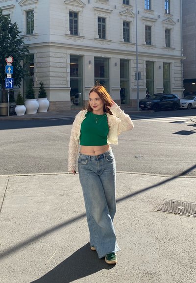 Modelo con un top corto verde, un cardigan texturizado crema y jeans de pierna ancha de color azul claro. Lleva zapatillas verdes; la luz del sol proyecta una sombra en el suelo.