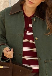 Vrouw in een groene jas over een maroon en wit gestreepte trui, die een bruine suède handtas vasthoudt, met een gouden ketting en oorbellen zichtbaar.
