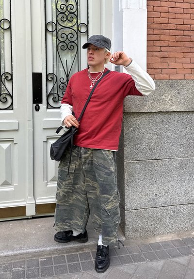 Camisa roja de manga corta colocada sobre una camiseta blanca de manga larga, pantalones holgados de camuflaje, zapatos negros, gorra gris y collares plateados.