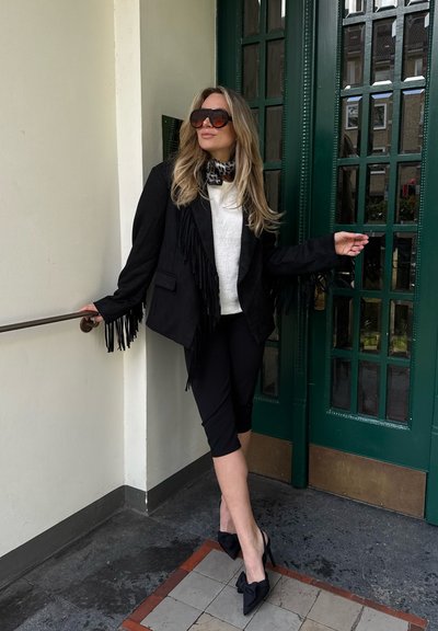Mujer con chaqueta negra con flecos, jersey blanco, pantalones cortos y tacones con lazo, de pie junto a una puerta verde con paneles de vidrio, lleva gafas de sol grandes y bufanda.
