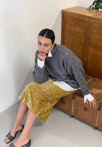 Siv prevelik pulover, pod njim bela srajca s ovratnikom, sijoča midi krilo z zlatimi sequini in črne natikače. Sedi na lesenem prtljažniku.