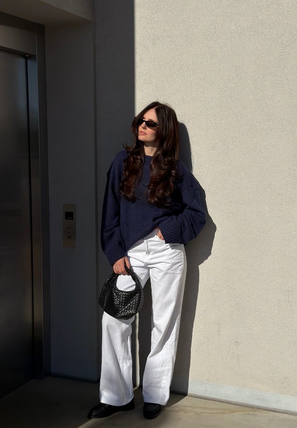 Mujer con cabello largo que lleva gafas de sol oscuras, suéter azul marino, pantalones anchos blancos, sosteniendo un bolso negro, parada junto al ascensor bajo la luz del sol.