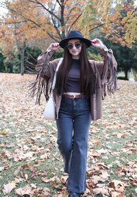Bruine jas met franjes, zwart topje, blauw hooggetailleerde jeans, zwarte hoed en ronde zonnebril. Vallende bladeren op de achtergrond.