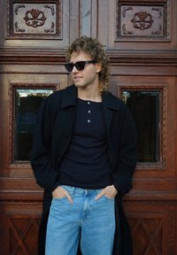 Musta ylipuku, laivastonsininen henley-paita, vaaleansiniset farkut ja aurinkolasit. Taustalla koristeltu puuovi, jossa on monimutkaisia kaiverruksia.