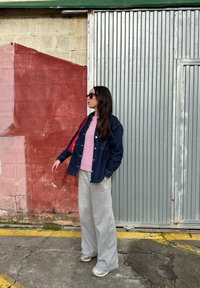 Chaqueta vaquera sobre una camisa de rayas rosas, combinada con pantalones deportivos grises y zapatillas plateadas. De fondo, una pared texturizada y pintada.