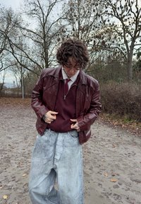 Maglione bordeaux con cerniera sotto una giacca di pelle color bordeaux; jeans grigi chiaro a gamba larga; scenario esterno con alberi e foglie cadute.