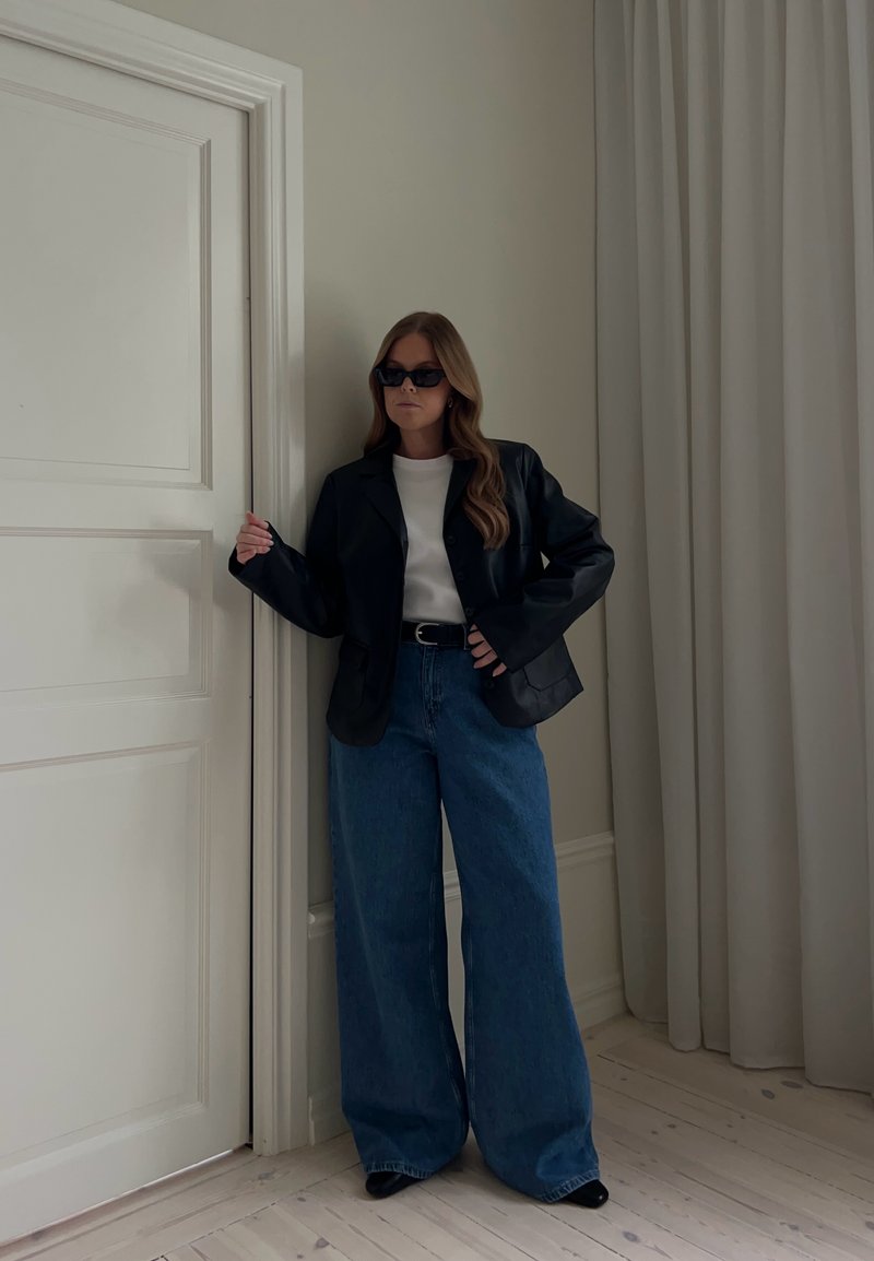 Femme portant une veste en cuir noire, un haut blanc, un jean bleu large, une ceinture noire et des lunettes de soleil, debout près d'une porte blanche et d'un rideau beige.