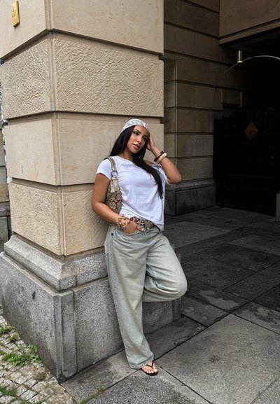 Camisa blanca de manga corta, pantalones vaqueros anchos de mezclilla clara, cinturón con estampado y una bolsa con estampado. Lleva chanclas y accesorios. Fondo de pared de piedra.