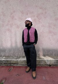 Μωβ τζιν γιλέκο πάνω από μαύρο τζάκετ, συνδυασμένο με μαύρα cargo παντελόνια και καφέ παπούτσια. Σε φόντο από υφασμάτινο ροζ τοίχο.