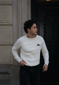 Λευκό πλεκτό πουλόβερ με ριγέ σχέδιο και στρογγυλή λαιμόκοψη, με πράσινο λογότυπο στο στήθος, φορεμένο με σκούρα τζιν μπροστά σε υφασμάτινο τοίχο.