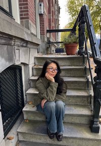 Une personne est assise sur des marches en pierre, portant une veste en daim verte, un jean évasé bleu et des chaussures marron, avec un pot de fleurs décoratif à proximité.