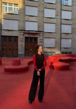 Chaleco negro sin mangas sobre una camiseta roja de manga larga, combinado con pantalones anchos negros. Superficie roja texturada con formas irregulares en el fondo.