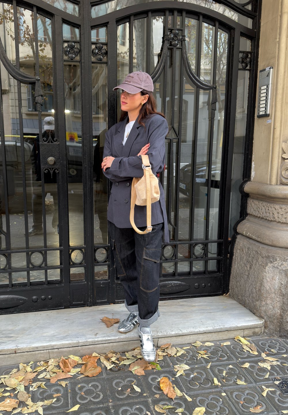 Femme en blazer gris, pantalon cargo noir, baskets argentées et casquette violette, debout les bras croisés près d'une grille en fer forgé noire.