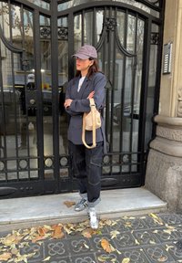 Femme en blazer gris, pantalon cargo noir, baskets argentées et casquette violette, debout les bras croisés près d'une grille en fer forgé noire.