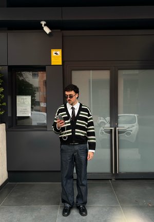 Cárdigan de rayas blancas y negras, camisa blanca, corbata negra, pantalones vaqueros. El usuario sostiene un teléfono inteligente con auriculares, de pie frente a puertas de vidrio.
