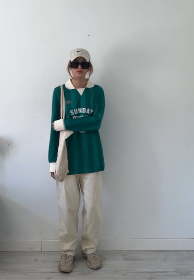 Jersey verde de manga larga con cuello blanco y diseño a rayas, combinado con pantalones crema y zapatillas beige. Una gorra ligera y gafas de sol completan el look.