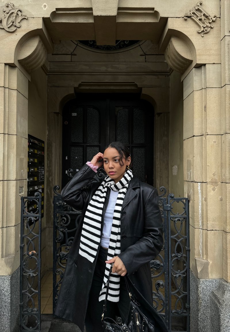 Manteau en cuir noir sur une chemise blanche, portant une écharpe rayée noir et blanc. Tient un sac noir, debout près d'une entrée en pierre ornée.