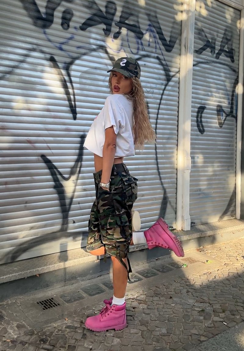 Καμουφλάζ cargo σορτς, λευκό κοντομάνικο μπλουζάκι, και φωτεινά ροζ μποτάκια με χοντρή σόλα. Το μοντέλο ποζάρει μπροστά σε έναν υφαντό, καλυμμένο με γκράφιτι τοίχο.