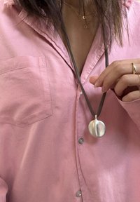 Pendente circolare in argento su una corda scura, indossato su una camicia button-up rosa. Il pendente ha una superficie liscia con un leggero riflesso.