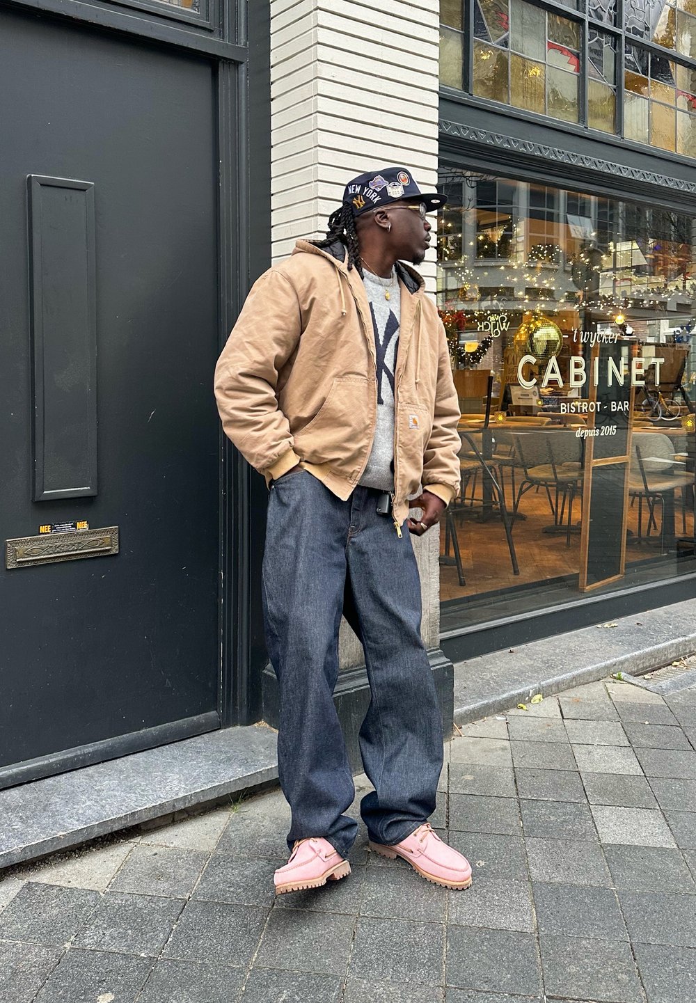 Homme portant une veste beige, un pull gris, un jean large foncé et des chaussures roses, debout sur le trottoir devant un café avec des fenêtres en verre et des décorations de fête.