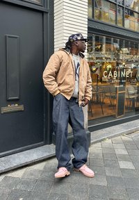 Homme portant une veste beige, un pull gris, un jean large foncé et des chaussures roses, debout sur le trottoir devant un café avec des fenêtres en verre et des décorations de fête.