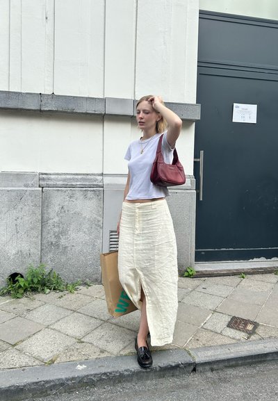 Camiseta blanca corta, falda crema texturizada con una abertura frontal, mocasines negros y un bolso burdeos. Bolsita de papel marrón con letras en azul verdoso.