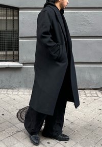 Jeune homme portant un long manteau noir, un pantalon noir et des chaussures noires, marchant sur un trottoir en ville près d'un mur gris et d'une fenêtre avec des barreaux.