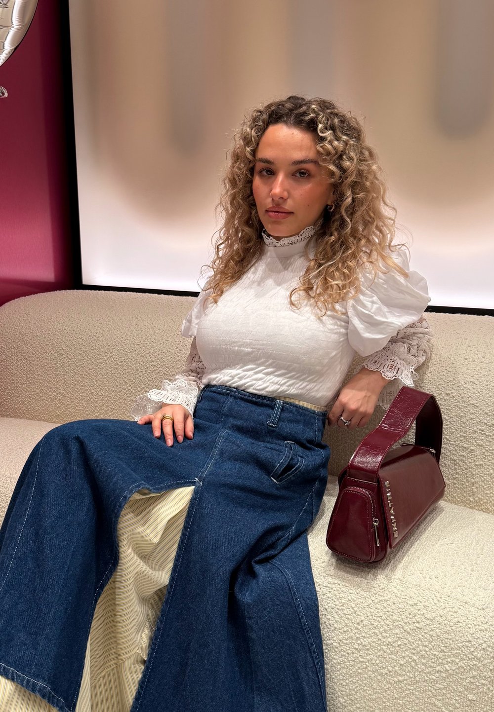 Blusa bianca con texture e maniche a sbuffo, abbinata a una gonna di denim con fodera a righe e una borsa bordeaux dal design angolare.