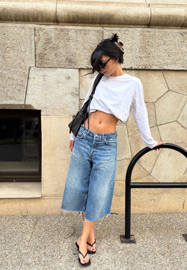 Mujer con camiseta corta blanca de manga larga, pantalones cortos amplios de mezclilla, gafas de sol negras y chanclas apoyada en un soporte metálico negro para bicicletas junto a una pared de piedra.