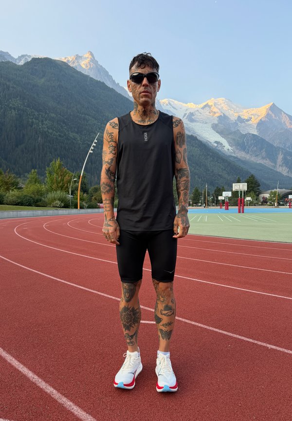 Man with full arm and leg tattoos wearing black athletic wear and sunglasses stands on red running track with mountains in the background.