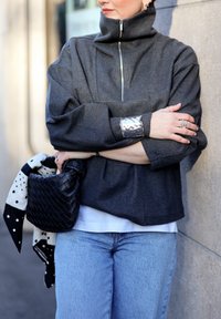 Personne portant un pull à fermeture éclair gris foncé, jean bleu clair, bracelet manchette en argent, tenant un sac à main noir tressé avec un foulard noir à pois blancs.