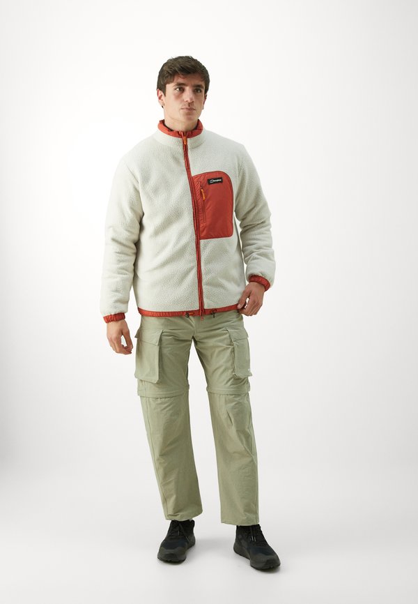Polartecová bunda v off-white barvě s červenou náprsní kapsou, oranžovým zipem a nastavitelnými manžetami; v kombinaci se světle zelenými cargo kalhotami a černými botami.
