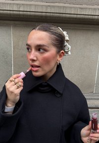 Manteau en laine noire avec col montant, tenant un gloss à lèvres rose et un produit à lèvres rouge foncé. Les accessoires comprennent des bagues et un chouchou pour cheveux.