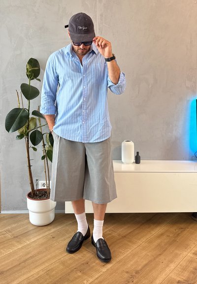 Camisa de botones a rayas azul claro, pantalones cortos grises, calcetines blancos, mocasines negros y una gorra gris, con una planta y una decoración mínima en el fondo.