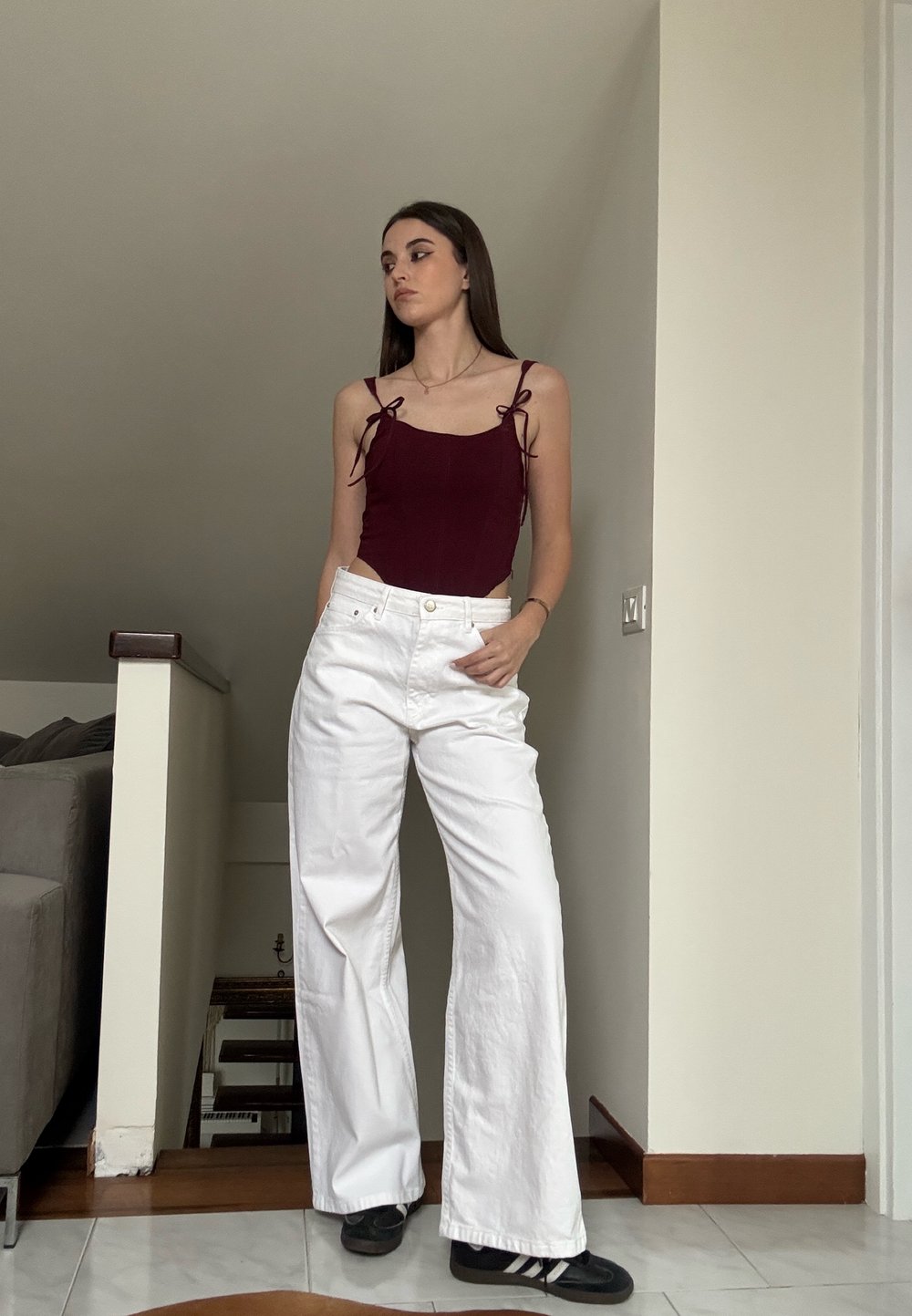 Bordeaux bodysuit met dunne bandjes en zijknoopjes, gecombineerd met witte wijde broek. Staande op een betegelde vloer in zwarte sneakers.