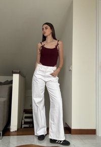 Bordeaux bodysuit met dunne bandjes en zijknoopjes, gecombineerd met witte wijde broek. Staande op een betegelde vloer in zwarte sneakers.