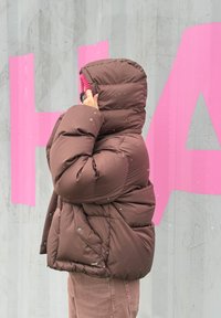 Brūna izolēta pūtuma jaka ar augstu apkakli un lielizmēra kapuci. Piedāvā gludu dizainu, vienmērīgu tekstūru un sprādzju pogas priekšpusē.