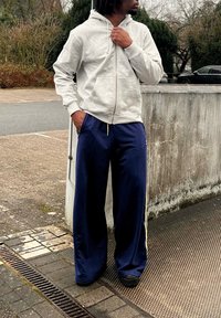 Homme portant un sweat à capuche zippé gris clair et un pantalon large bleu marine, debout en plein air avec une main dans la poche et l'autre près du cou.