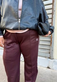 Jaqueta bomber em couro preto com um fecho prateado, combinada com calças jogger bordô com riscas brancas e um logótipo da Adidas. Segurando uma clutch preta trançada.