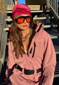 Rosa wasserdichte Jacke mit schwarzen Akzenten, ausgestattet mit einem Gürtel, Reißverschlusstaschen und Logo. Kombiniert mit übergroßen orangefarbenen Sonnenbrillen und einem rosa Kopftuch.