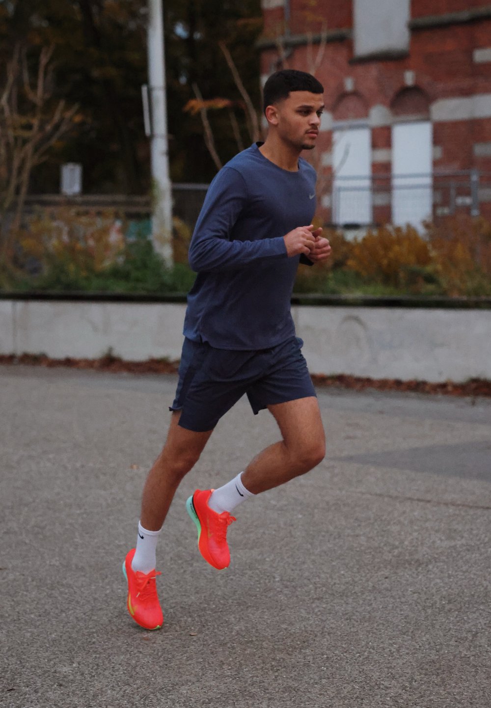 Athlète en maillot et short de sport bleu marine courant sur le trottoir. Chaussures rose fluo vif avec des accents verts. En arrière-plan, des arbres et un bâtiment en briques.