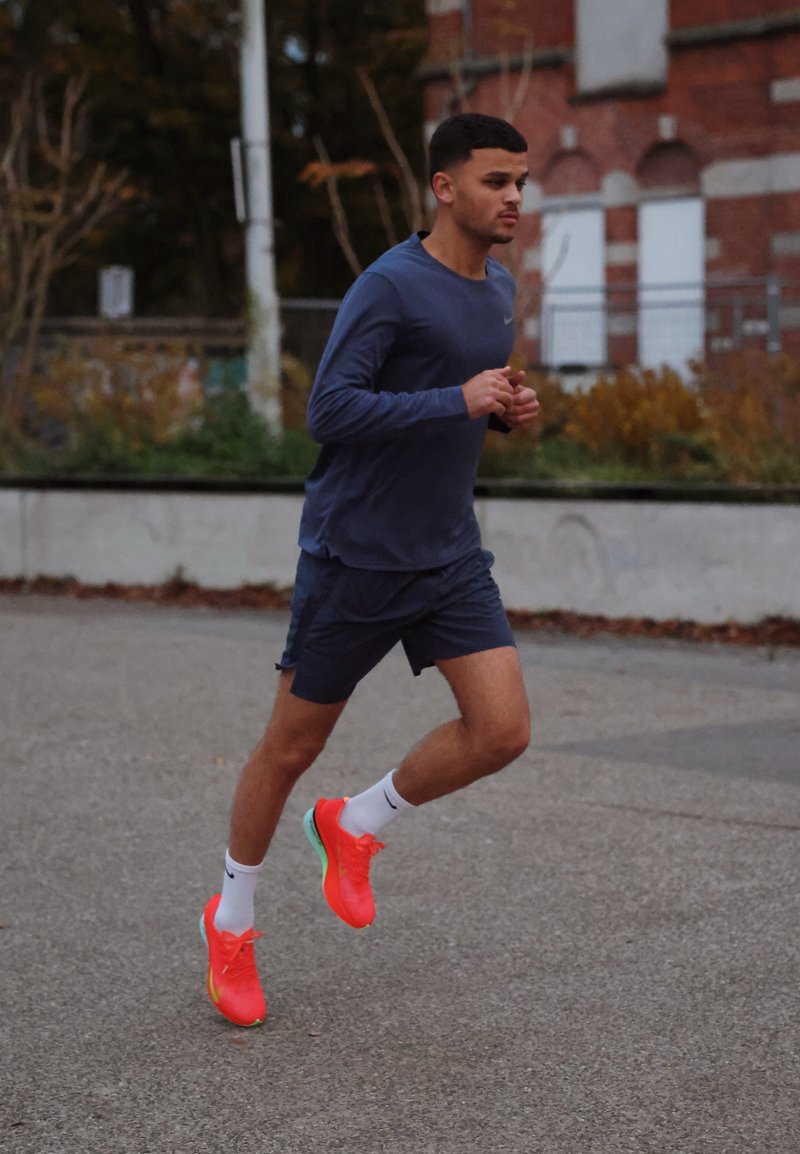 Athlète portant un t-shirt et un short de sport bleu marine courant sur un trottoir. Chaussures rose néon vif avec des accents verts. Arrière-plan comprenant des arbres et un bâtiment en briques.